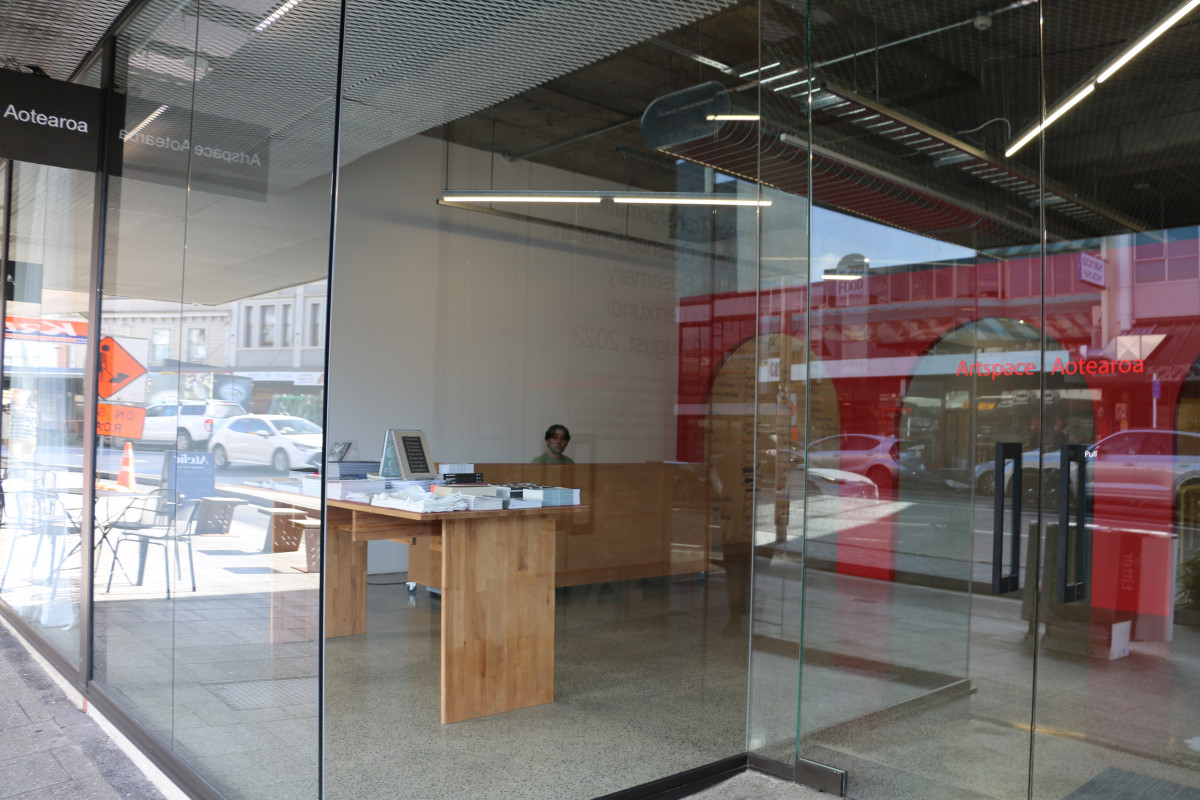 A fully glazed facade is visible. There are two glass doors with red writing on it that say Artspace Aotearoa. There is a books table in the window. A Kaitaki Gallery Host can be seen through the windows in our welcome area. Lights on in the gallery indicate the gallery is open.