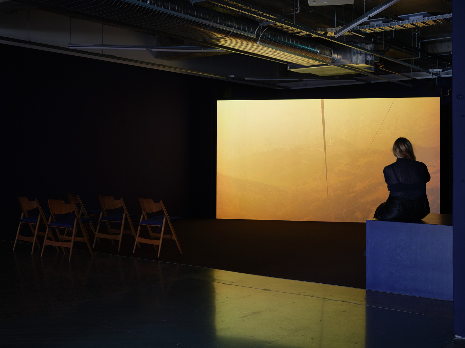 A large screen with foggy orange light is positioned at the back fo a room with someone sitting watching the film on a platform and seating to the right.