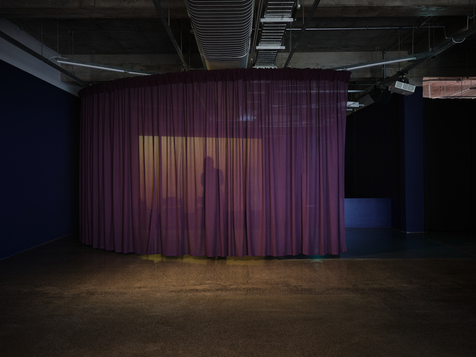 A large transparent curtain is hung form the ceiling, orange light from a screen shines through the fabric.