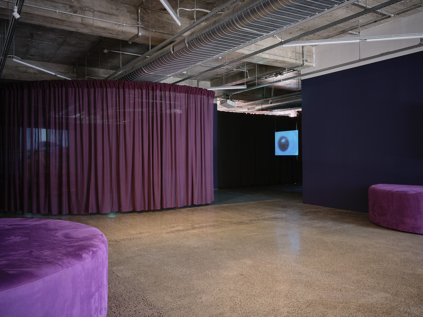 A gallery space has two semi-circle seats sitting against the wall and a large curtain drawn across the back of the space. A screen can be seen to the side of the curtain.