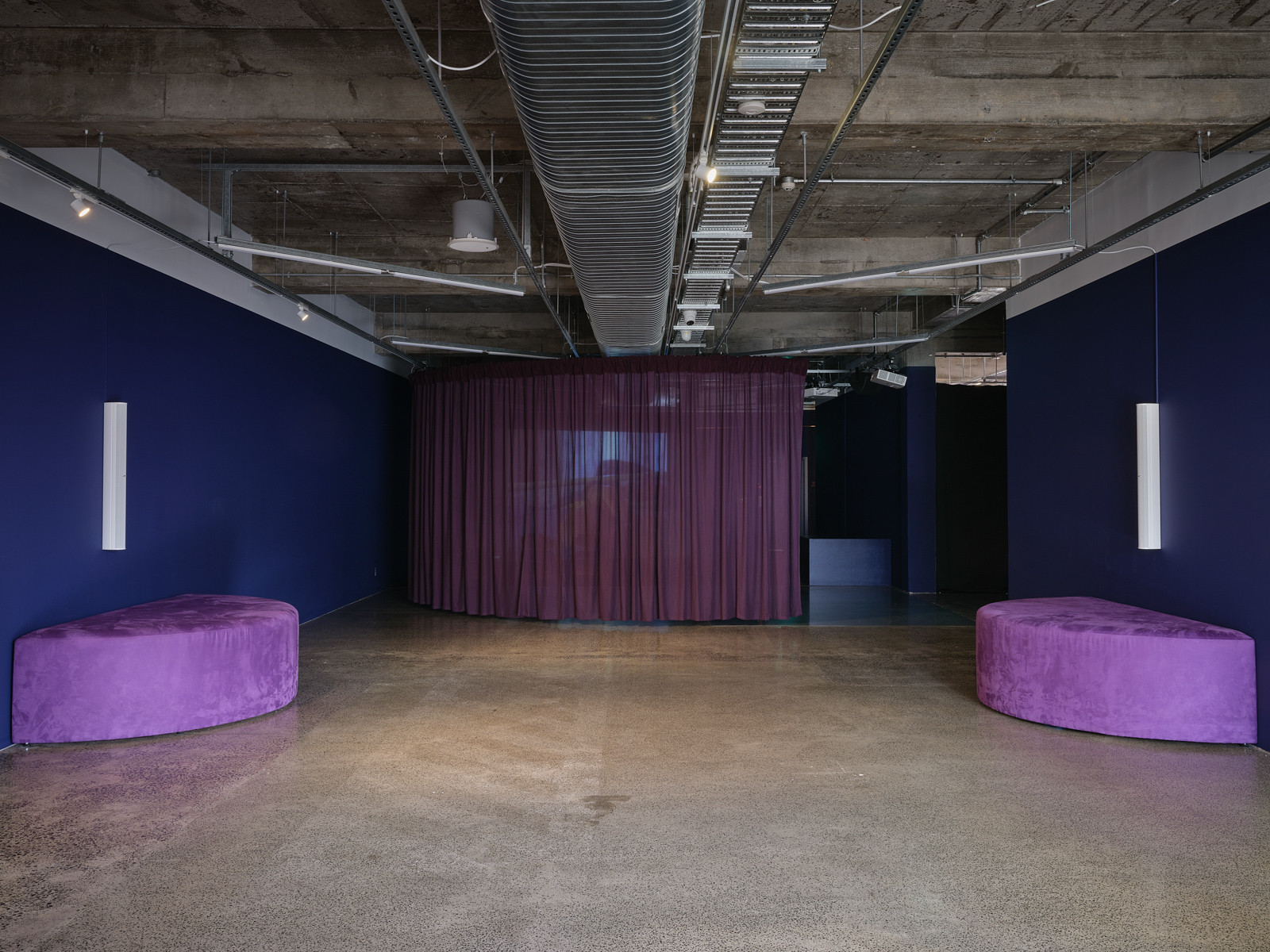 A gallery space has two semi-circle seats sitting against the wall and a large curtain drawn across the back of the space.