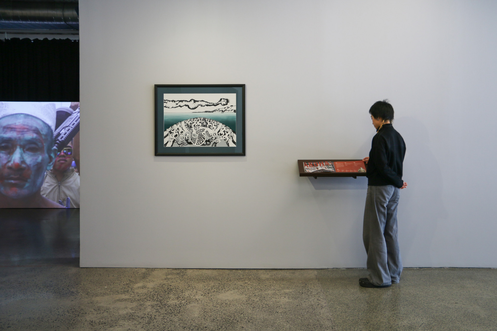 A framed print and shelf with a publication are hung on a gallery wall, with a projection in the background and a person is looking at the publication.