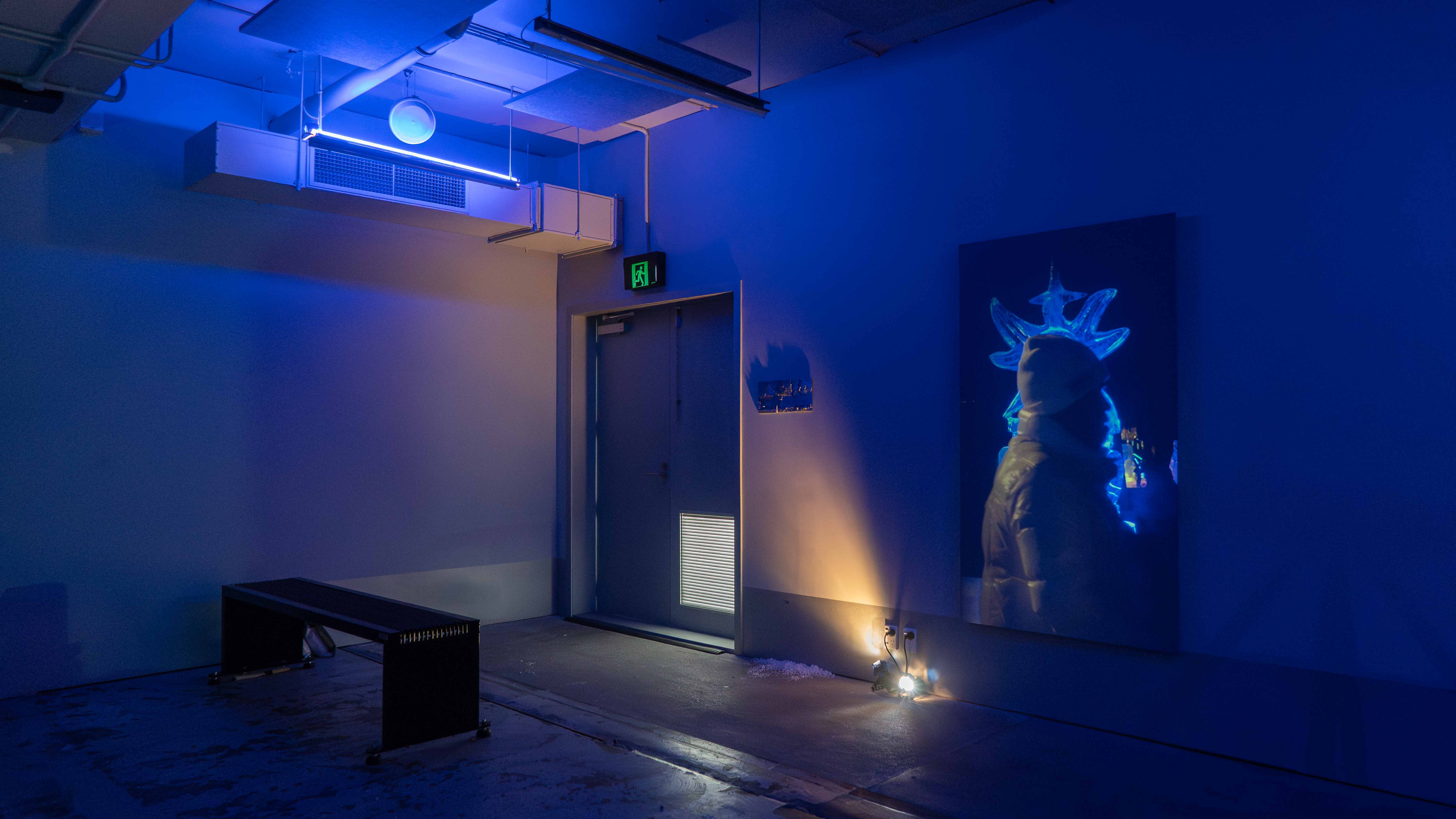 In a dark industrial space, a black metal bench is set near the entrance and a projection showing people walking pass a 2 meters tall ice sculpture