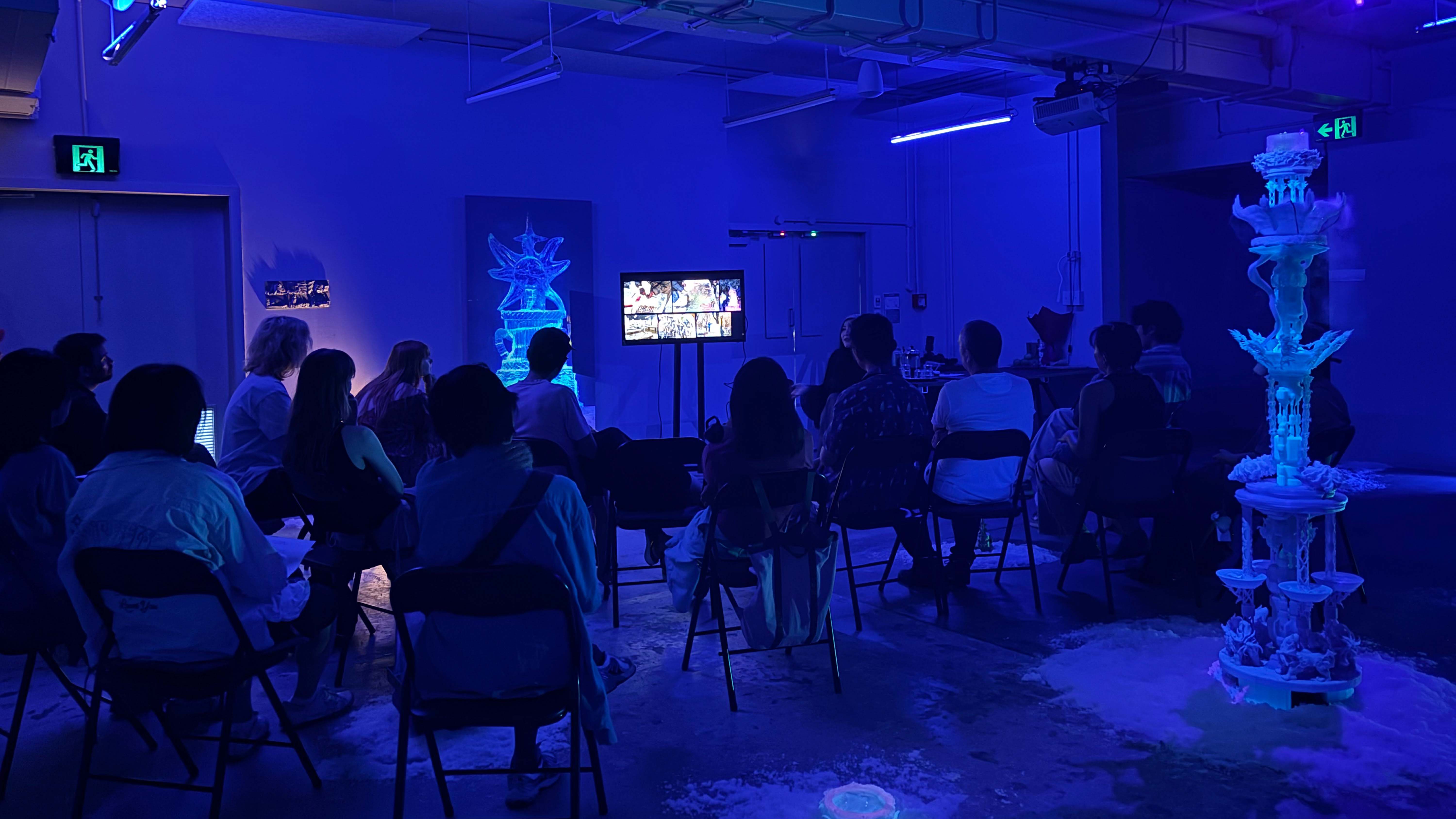 20 people sitting on chairs facing a presenter talking in a dark industrial space with art works