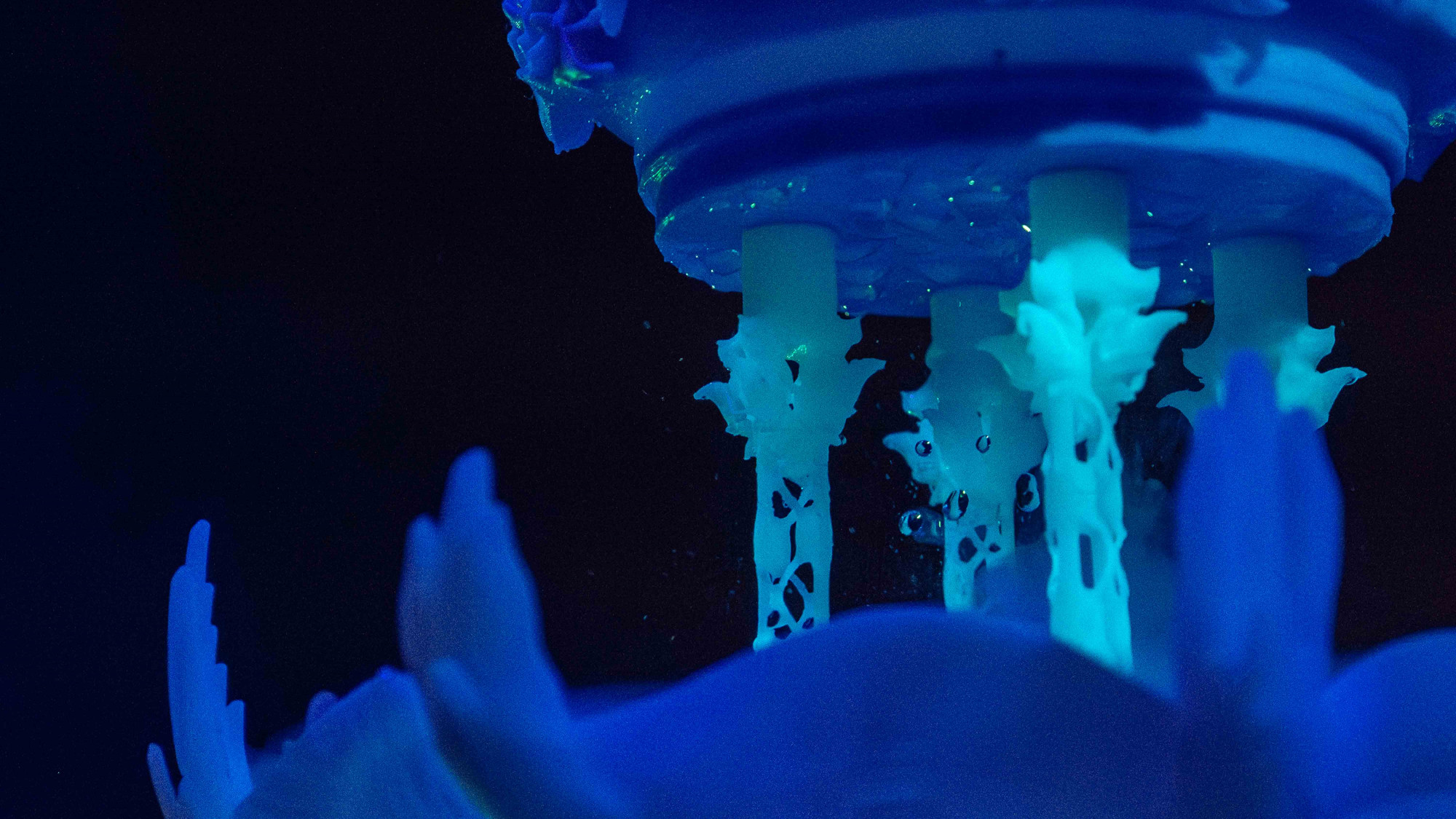 An illuminated sculpture in glow in dark green and blue, water drops are floating with mist inside its structure