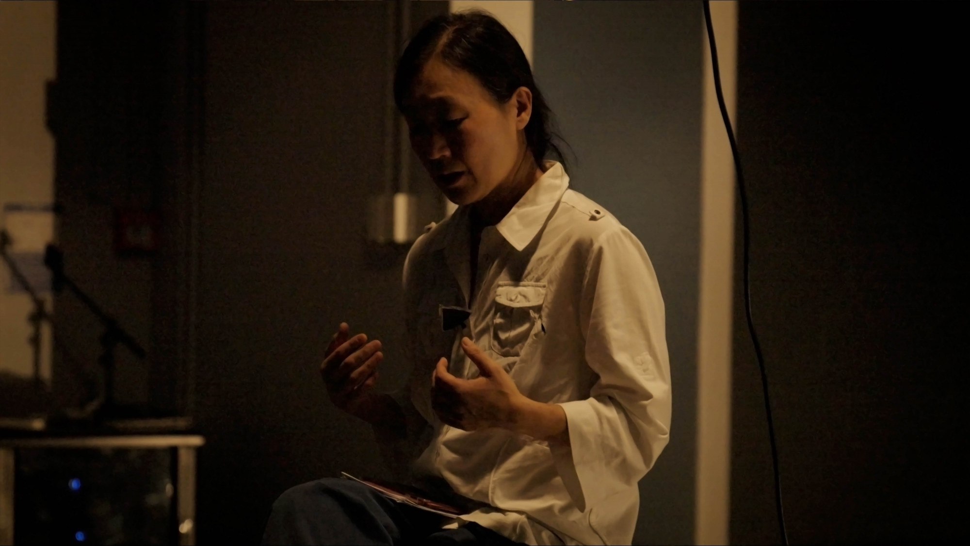 In a dark room with warm yellow light, a woman in white shirt talking