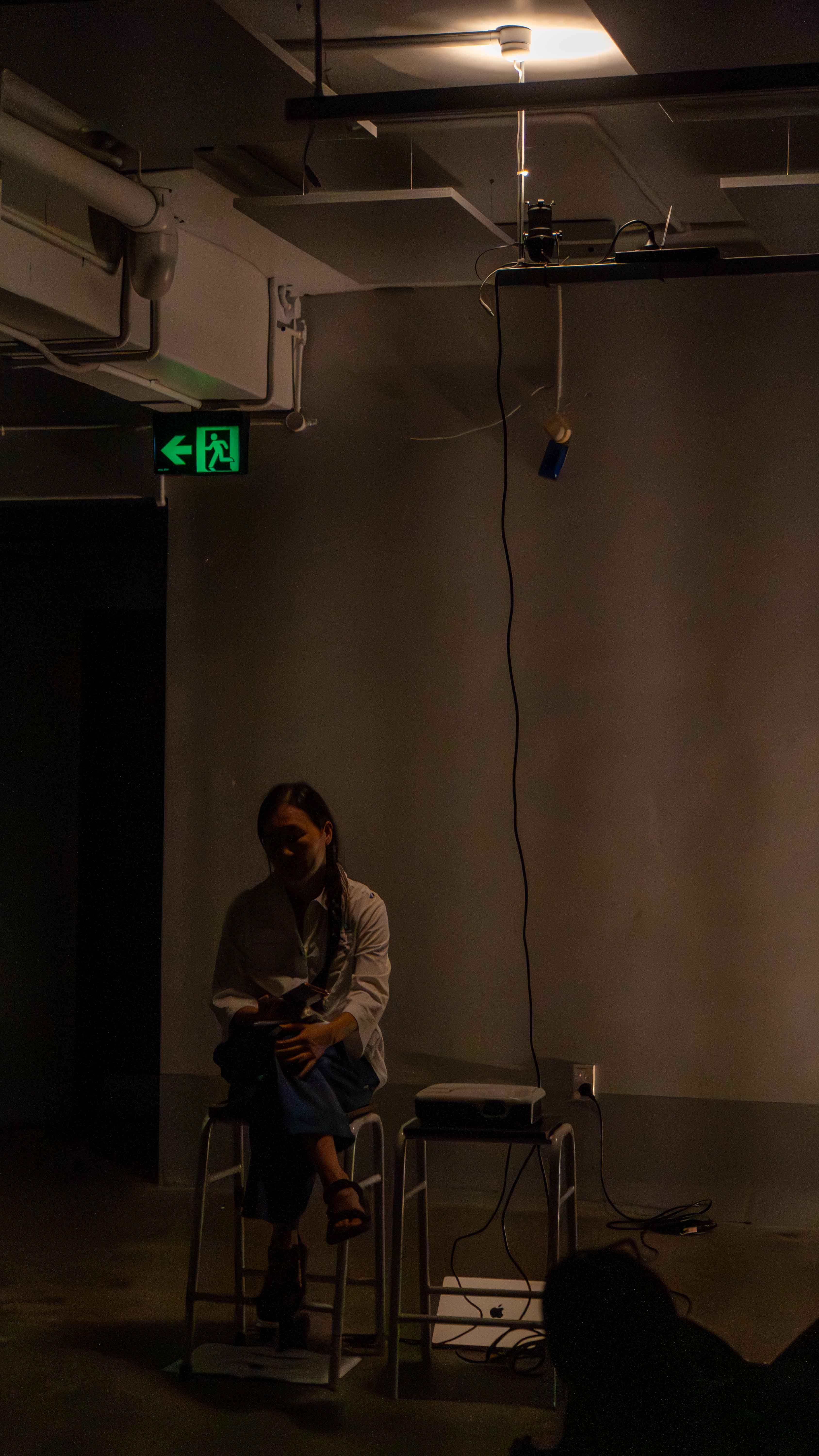 In a dark room with warm yellow light, a woman in white shirt sitting on a stool cross-legged