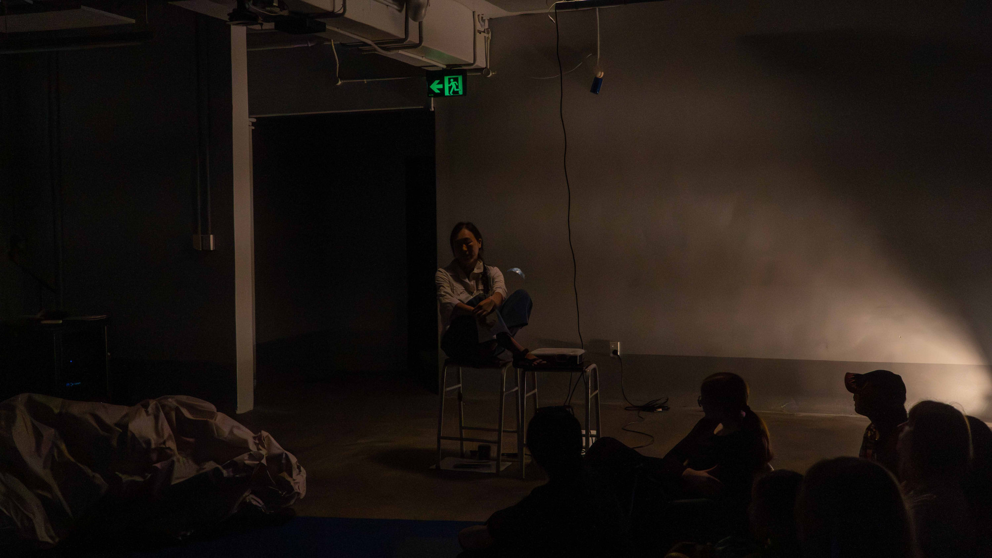 In a dark room with warm yellow light, a woman in white shirt sitting on a stool cross-legged