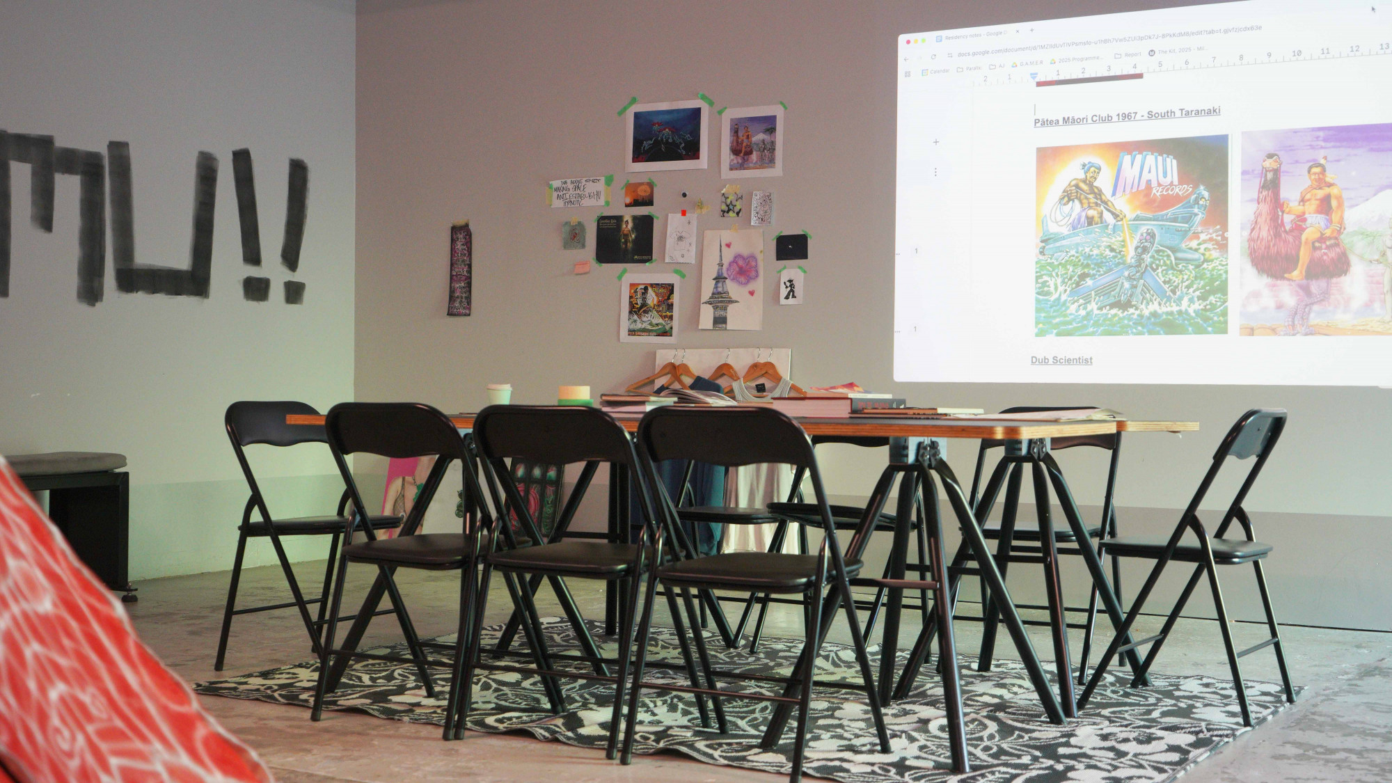 a table with books surrounded by chairs, a projection on the wall displaying covers from Pātea Māori Club record.