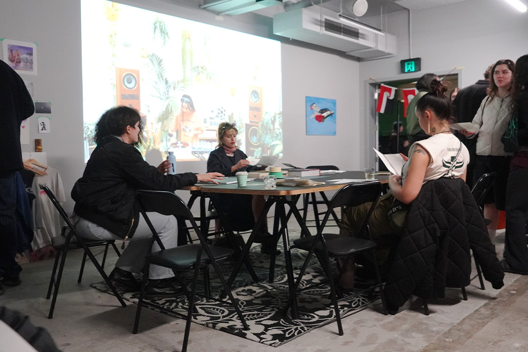 Four people sitting and reading around a table with books next to a crowd of  people standing and talking.