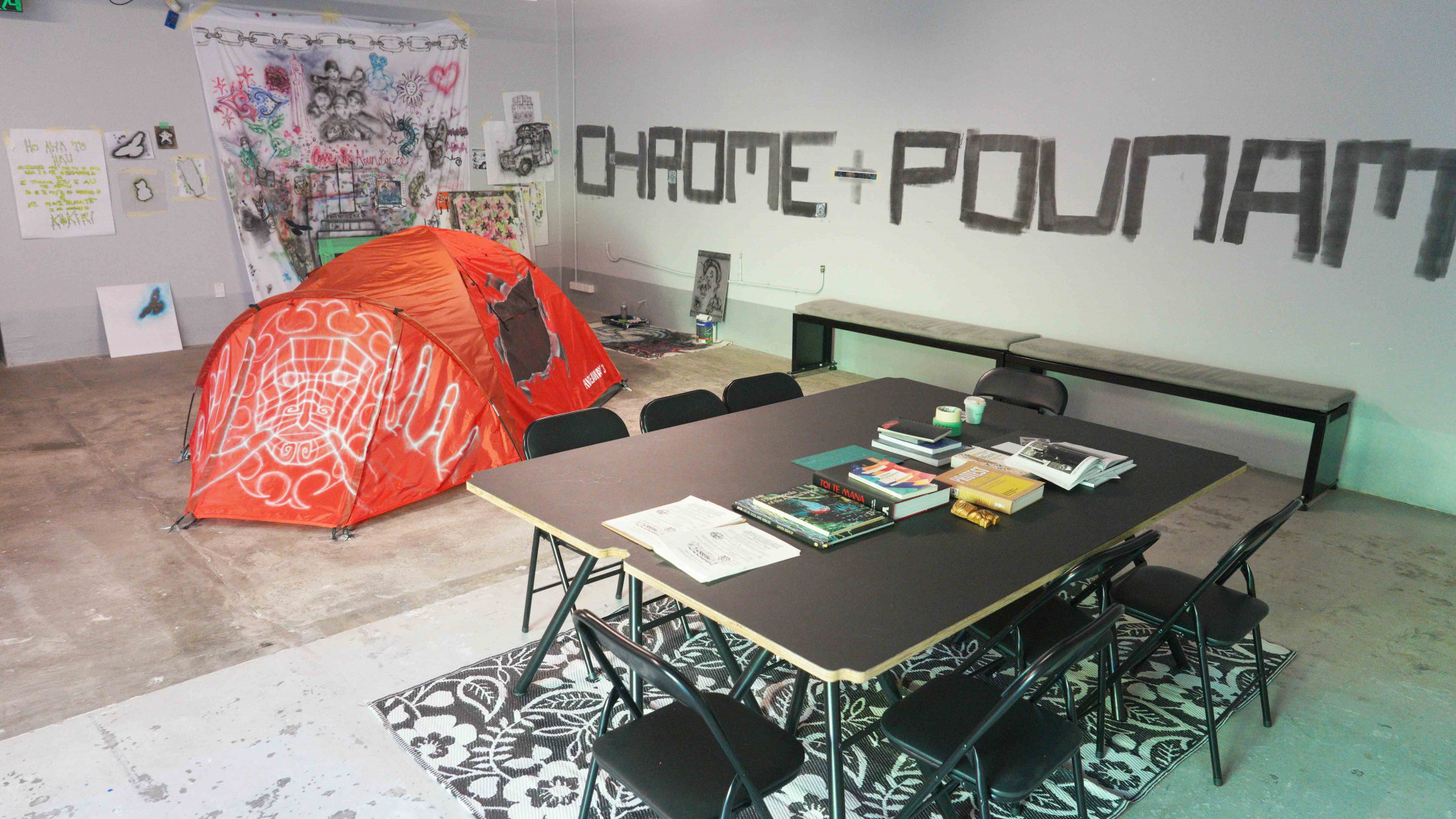 A table with many books and an air brush painted orange tent and drop cloth in the background. The sentence Chrome + pounamu is written on the back wall.